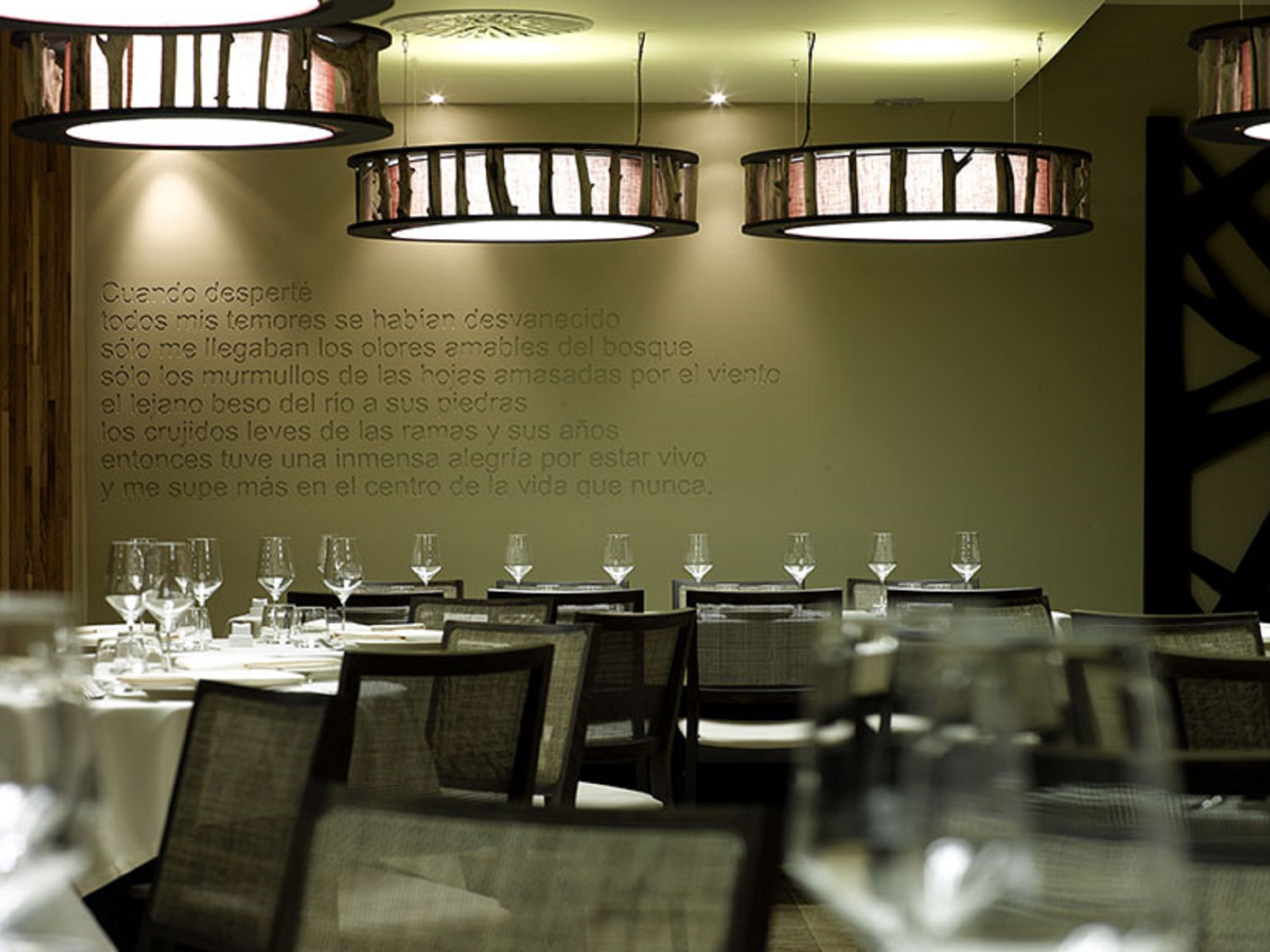 A restaurant interior with multiple tables set for dining, overhead lighting fixtures, and a wall with a poem in Spanish.