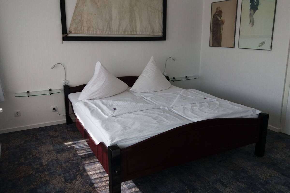 A neatly made bed with white linens and two pillows, a dark wooden headboard, a glass shelf with a small item on it, and a framed artwork above the bed.
