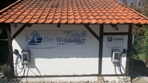 Um edifício com telhado em tijolos e uma placa com a inscrição 'Der Waldsegler Hotel'. Há estações de carregamento para veículos elétricos na lateral.
