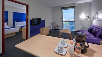 A hotel room with a bed, a desk with a coffee maker, a chair, a purple sofa, and a large window with a city view.