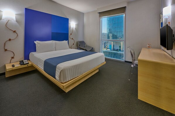 A hotel room with a bed, bedside table, chair, desk, TV, and a window with a view of the city.