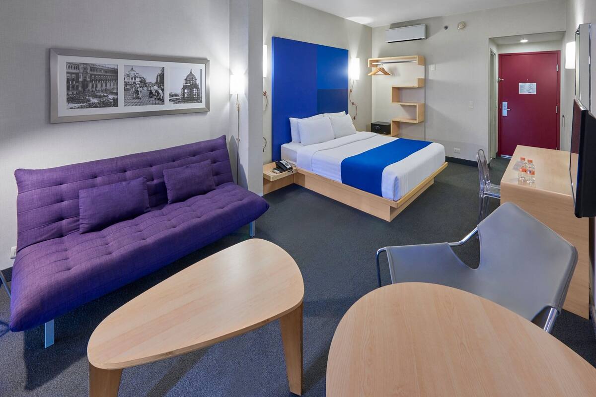 Habitación de hotel con sofá morado, cama con cabecera azul, una mesa de madera, una silla y un escritorio con televisor.