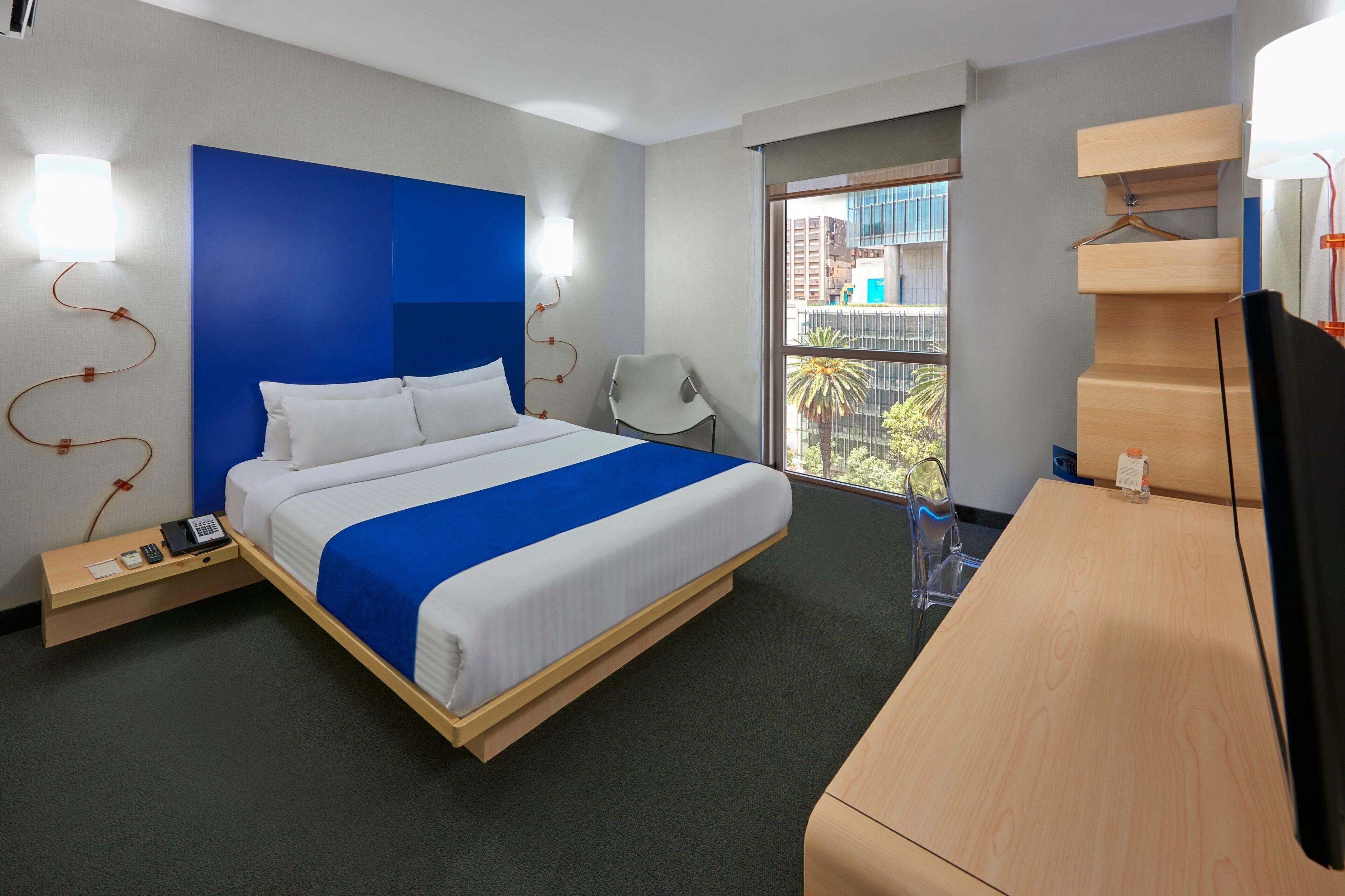 A modern hotel room with a large bed, bedside tables, a desk, and a view of the city through the window.