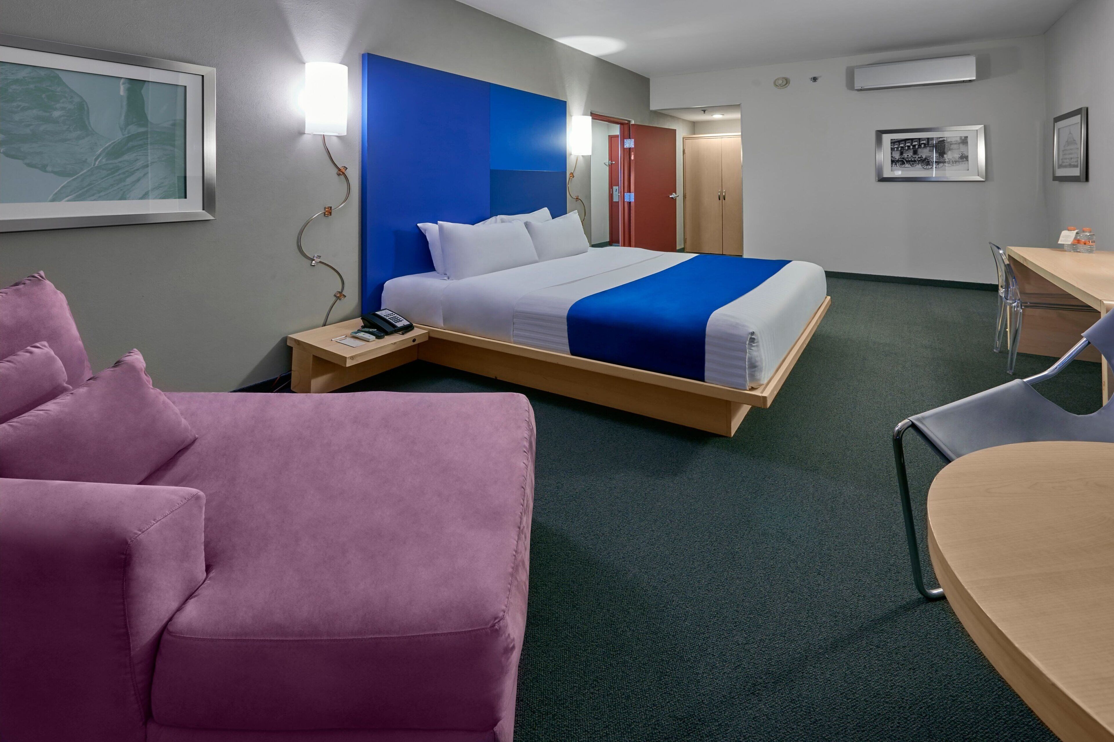 Una habitación de hotel con cama, escritorio, silla, sofá rosa y un cuadro enmarcado en la pared.