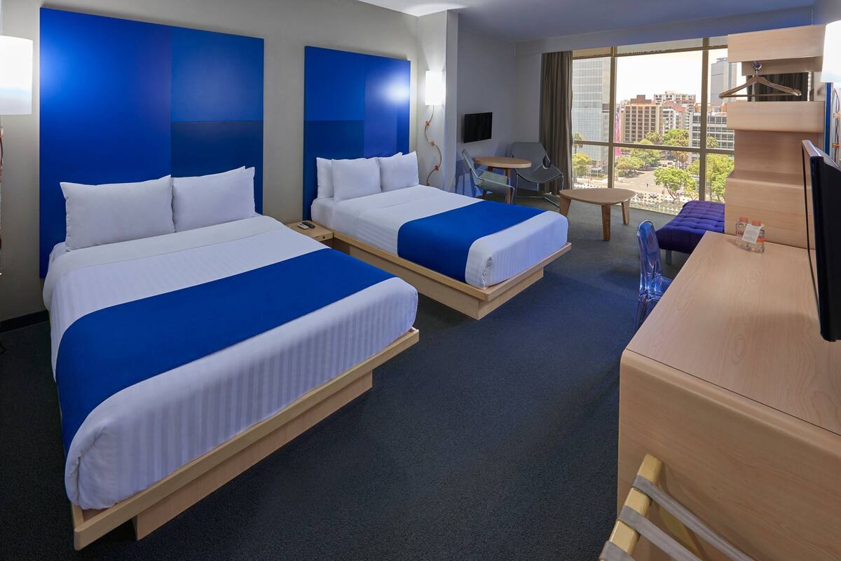 Habitación de hotel con dos camas, un escritorio, una silla, un televisor y vistas a la ciudad.