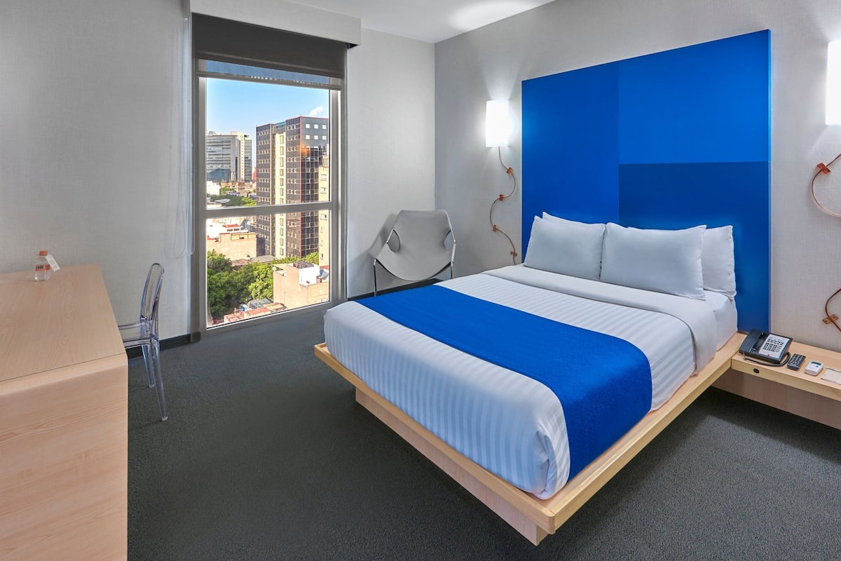 Habitación de hotel con cama, una silla, escritorio y ventana con vista a la ciudad.