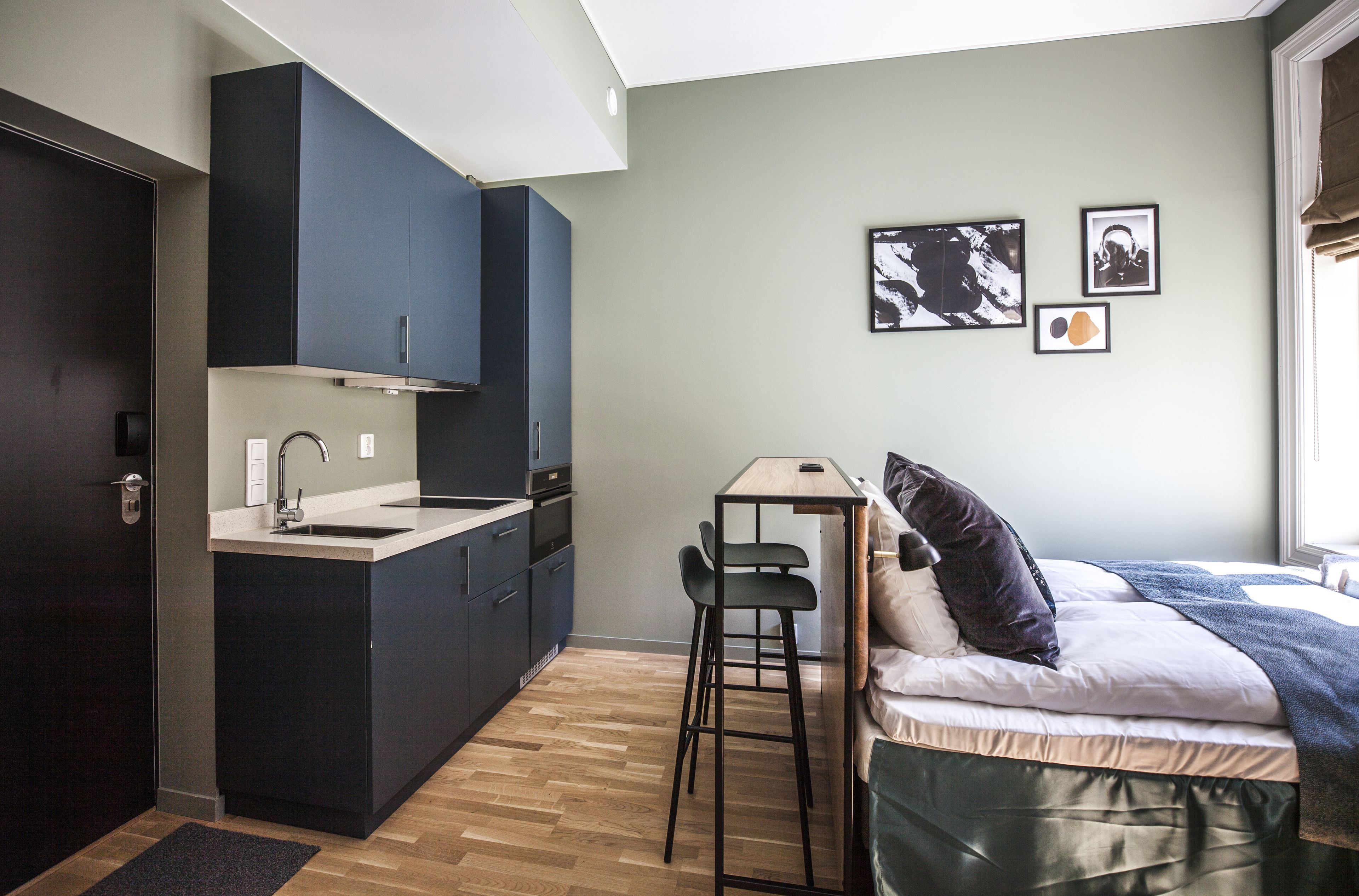 A compact kitchen with dark cabinets, a sink, and a small dining area with stools. There is a bed with a pillow and a blanket, and a window with curtains.