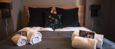 A neatly made bed with a dark grey bedspread, two pillows, and rolled towels.