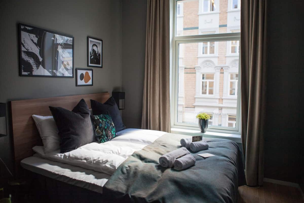 Een netjes opgemaakt bed met kussens, een nachtkastje met een lamp, en een raam met gordijnen.