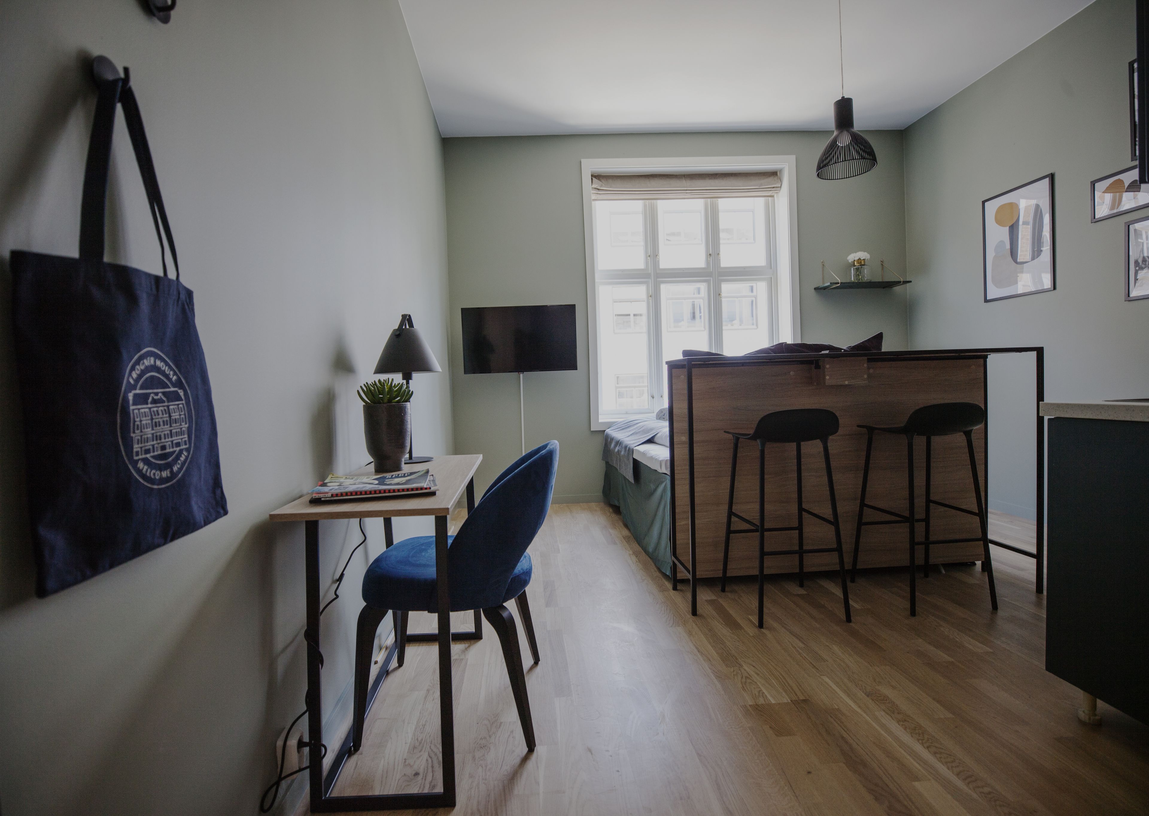 A compact living space with a kitchenette, a dining area with a bar stool, a small table with a lamp, and a large window providing natural light.