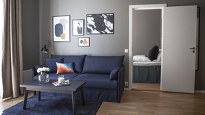 Un salon moderne avec un canapé bleu foncé, une table basse ornée d’un vase et d’une bougie, et une porte donnant sur une chambre à coucher équipée d’un lit et d’une table de chevet.