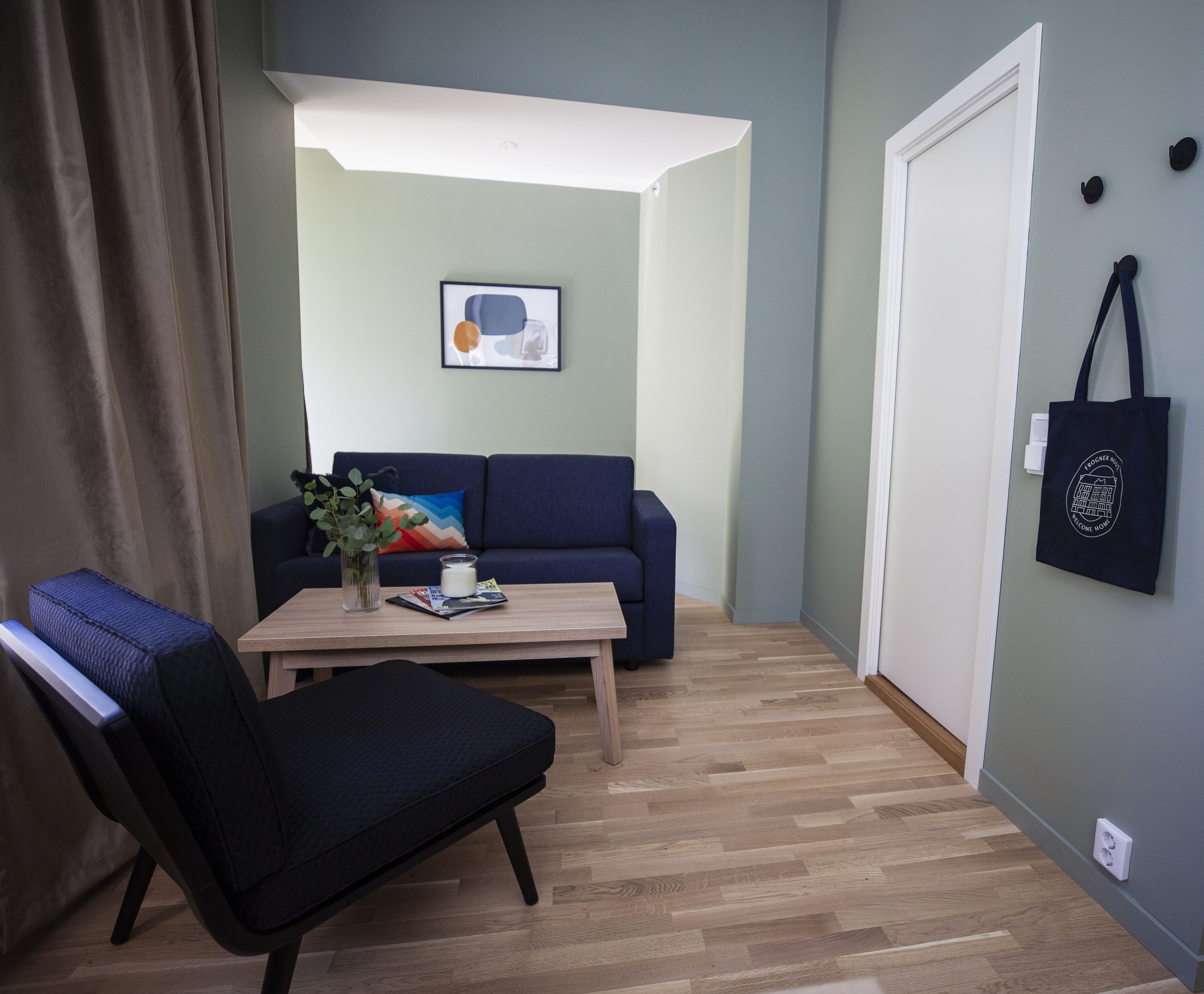 Ein modernes Wohnzimmer mit einem blauen Sofa, einem Holztisch und einem schwarzen Sessel. Ein gerahmtes Bild hängt an der Wand, und eine schwarze Tasche hängt an der Tür.
