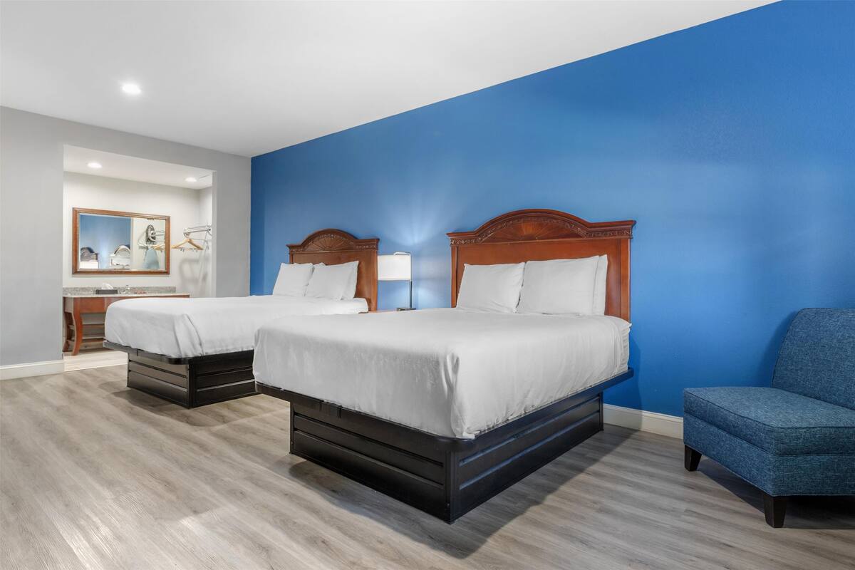 A hotel room with two beds, a blue accent wall, a wooden floor, and a chair.