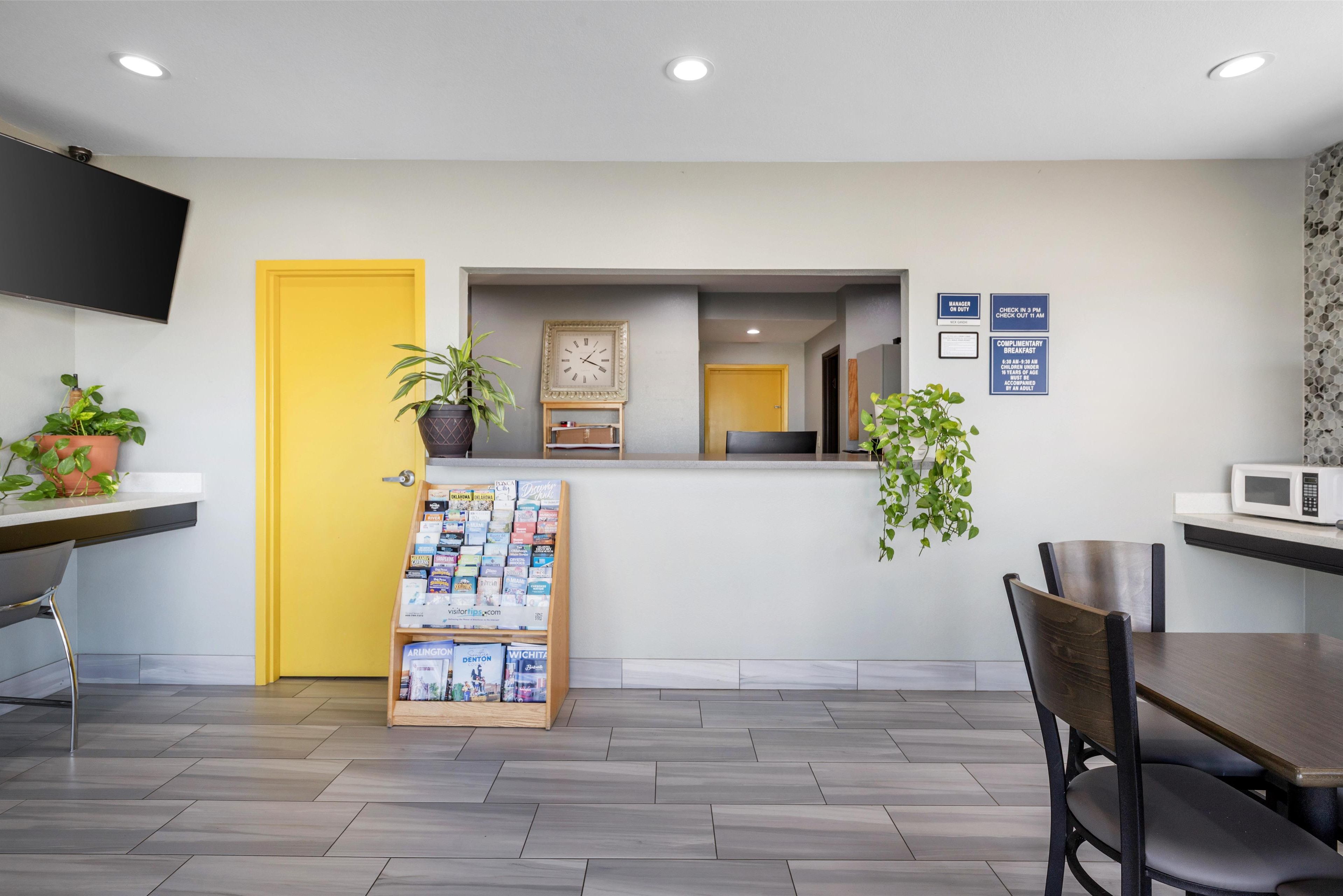 A modern hotel lobby with a yellow door, a reception desk, a clock, and a wall-mounted TV.