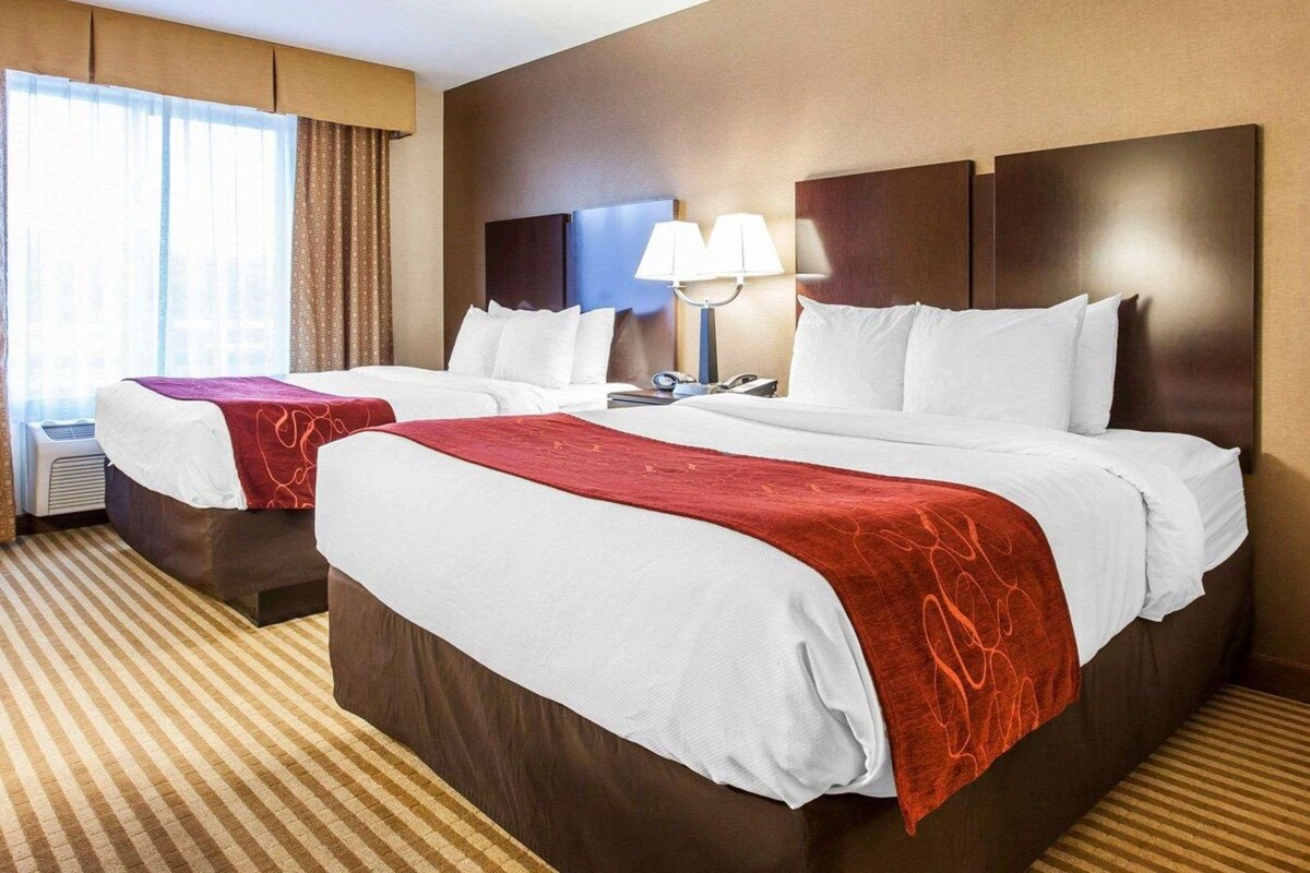 A hotel room with two beds, a large window with curtains, a bedside table with a lamp, and a striped carpet.