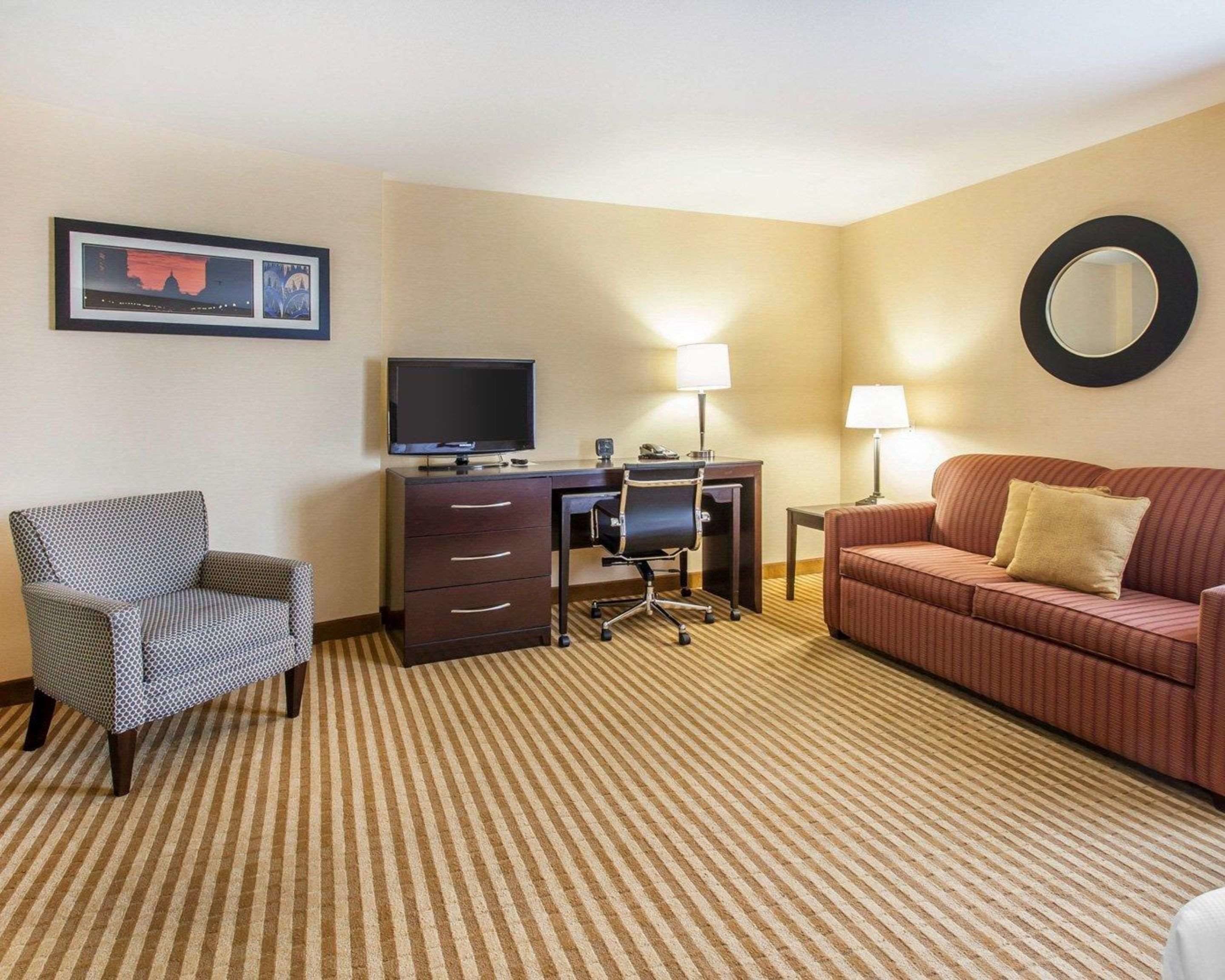 A hotel room with a striped carpet, a desk with a computer and a TV, a chair, a sofa, and two lamps.
