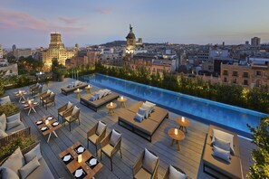 A rooftop pool area with lounge seating and tables, surrounded by city buildings.