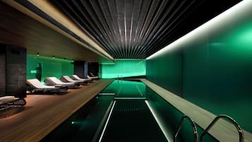 A modern spa with a long pool, wooden loungers, and a green-lit ceiling.