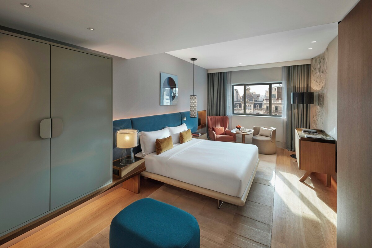Habitación de hotel con una cama grande, un escritorio, una silla, una mesita y una ventana con vista a la ciudad.