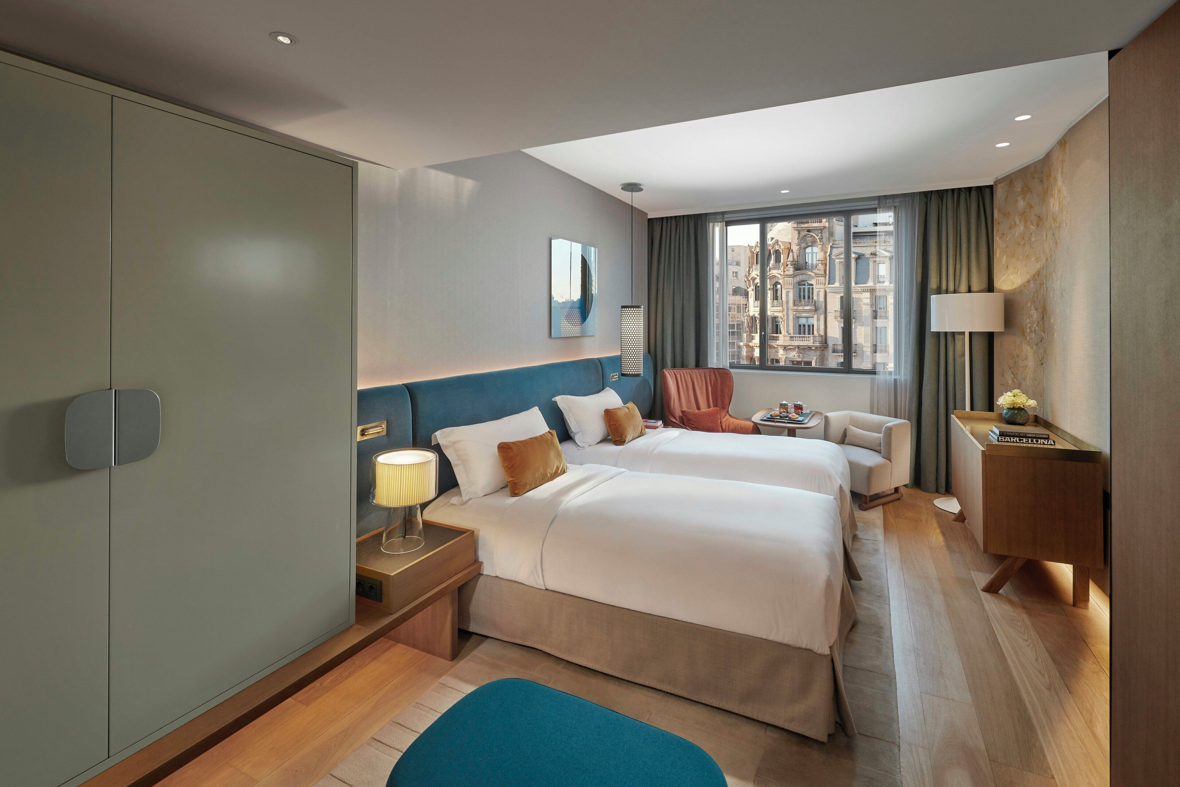 A hotel room with two beds, a desk, a lamp, and a view of the city.