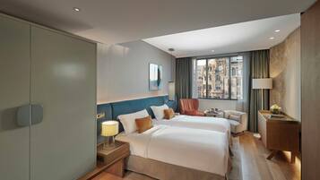 A hotel room with two beds, a desk, a lamp, and a view of the city.