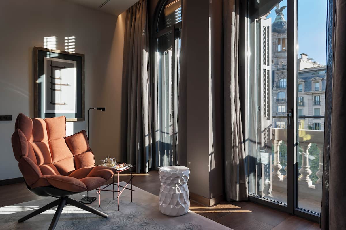 A modern living room with a large window, a cushioned chair, a small table, and a decorative stool.