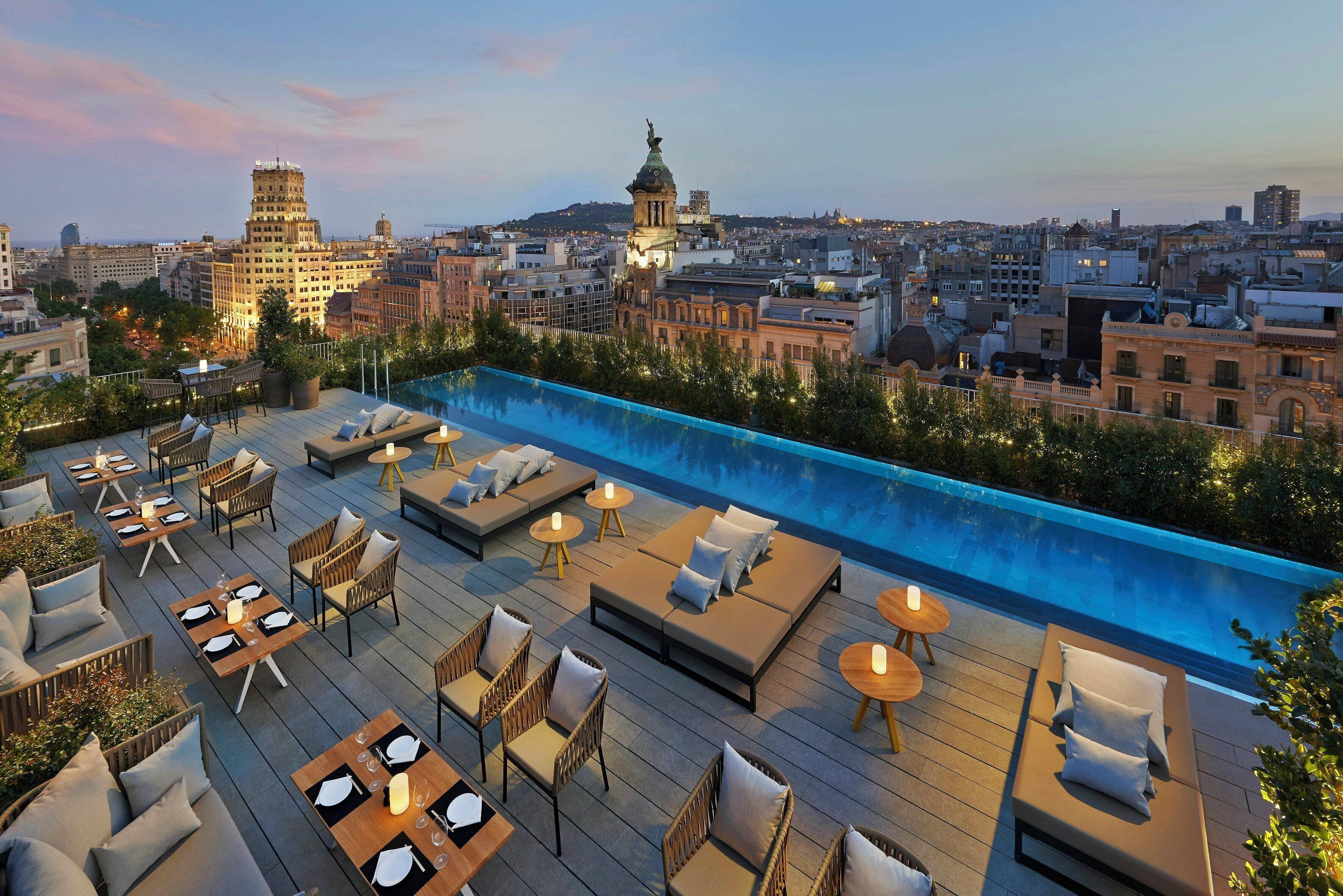 Zona de piscina en la azotea con sillones de descanso y mesas, con vistas a un paisaje urbano con edificios históricos.