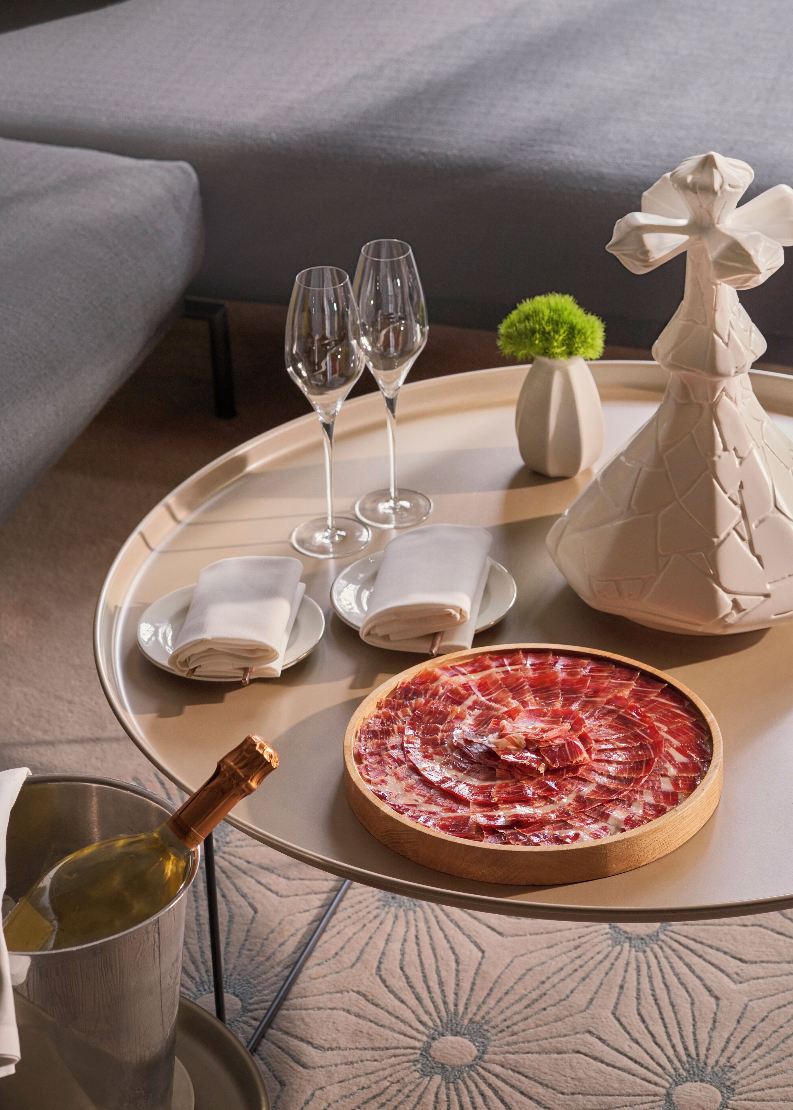 A dining table with a plate of cured meat, two glasses, a bottle of wine, and a decorative vase.