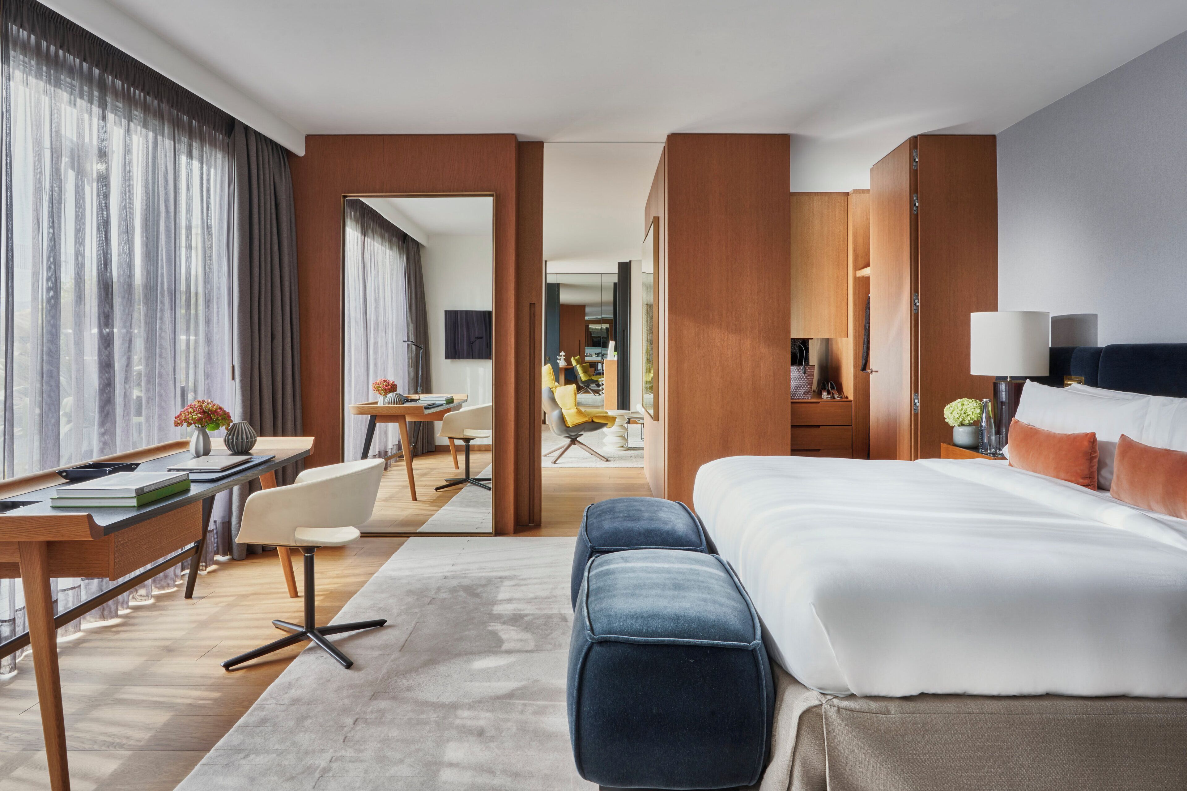 A modern hotel room with a large bed, a desk with a chair, a small table, and a sofa. There is a mirror on the wall and a window with curtains.