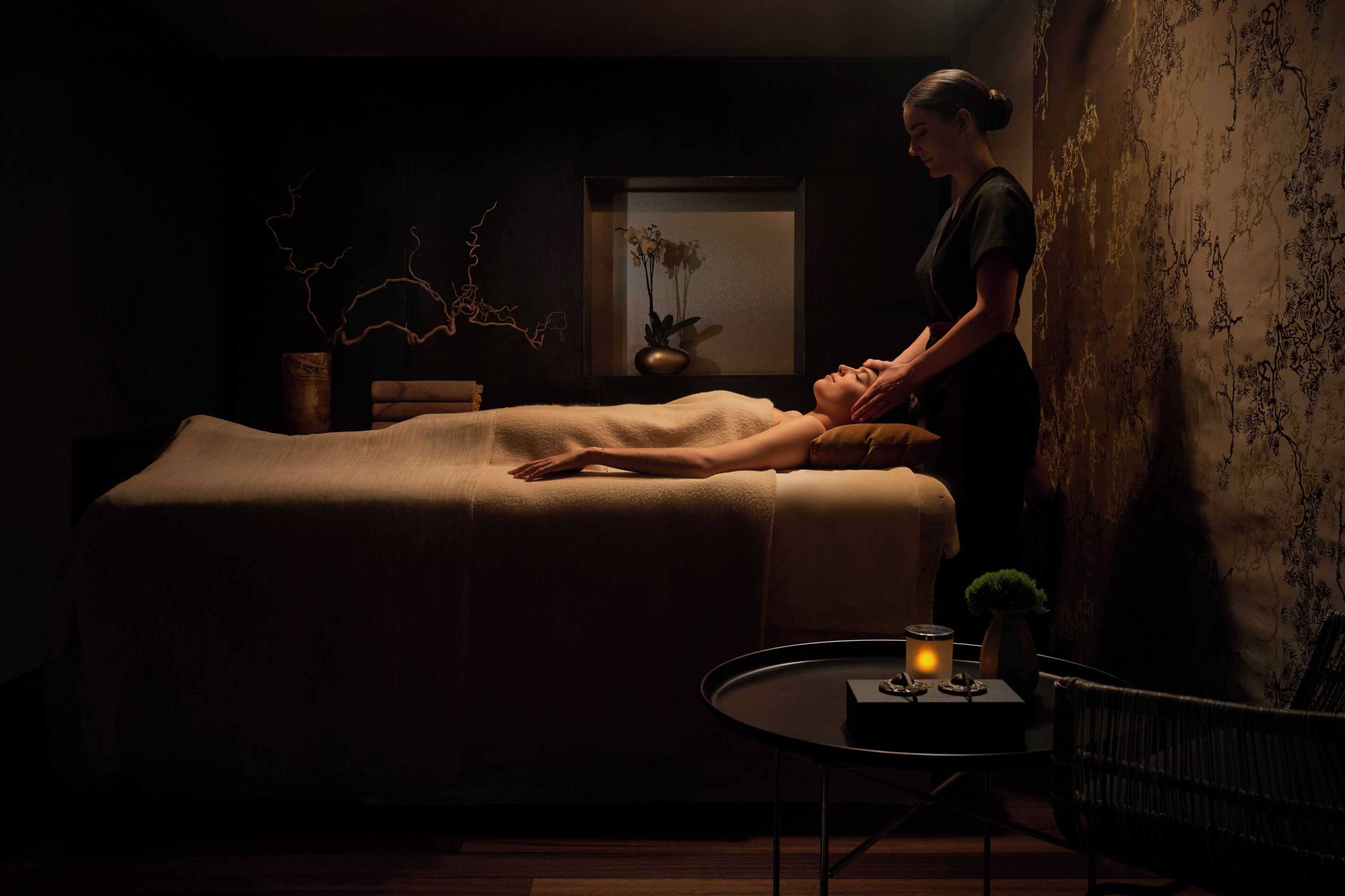 A dimly lit room with a massage table, a small table with a candle, and a wall with a decorative pattern.