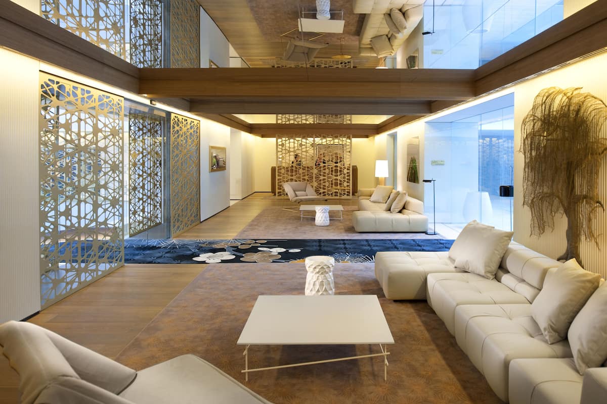 A spacious living area with a large white sofa, a white coffee table, and a patterned rug. There is a decorative wall with intricate designs and a large plant on the right.