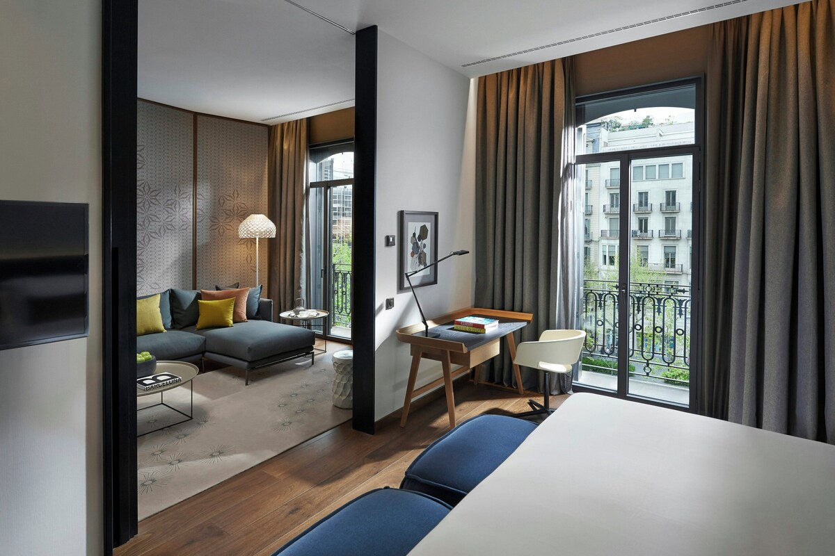 Una habitación de hotel moderna con sofá, mesa de comedor, cama y ventanas grandes con cortinas.