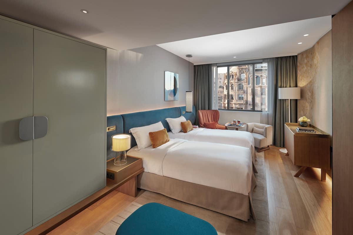 Quarto de hotel com duas camas, uma escrivaninha, uma cadeira, um abajur e vista da cidade por grandes janelas.