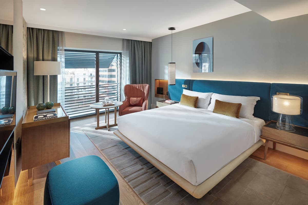 Quarto de hotel moderno com uma cama grande, uma escrivaninha, uma cadeira e vista para a cidade através de amplas janelas.