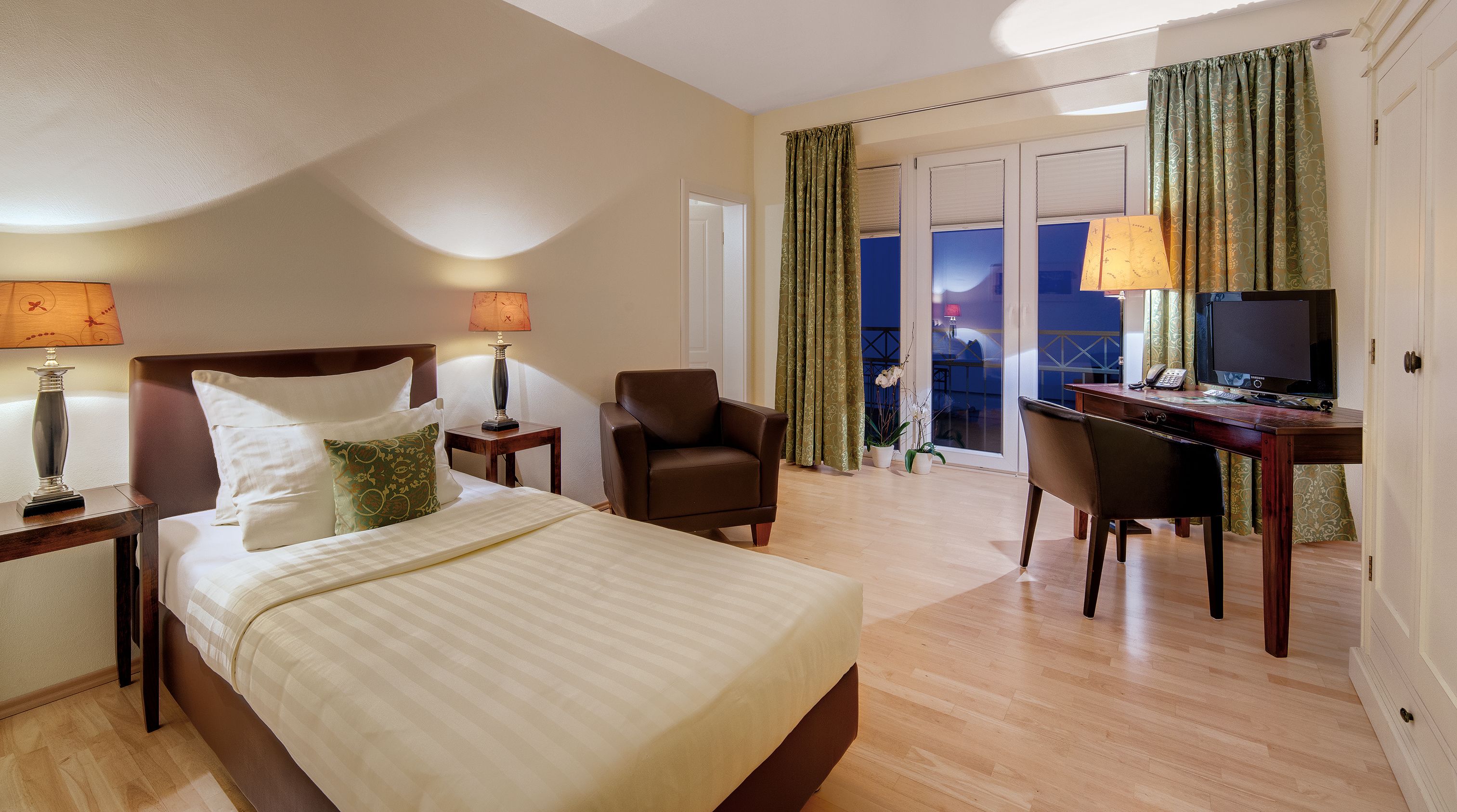 A hotel room with a bed, a desk, a chair, a TV, and a balcony with curtains.