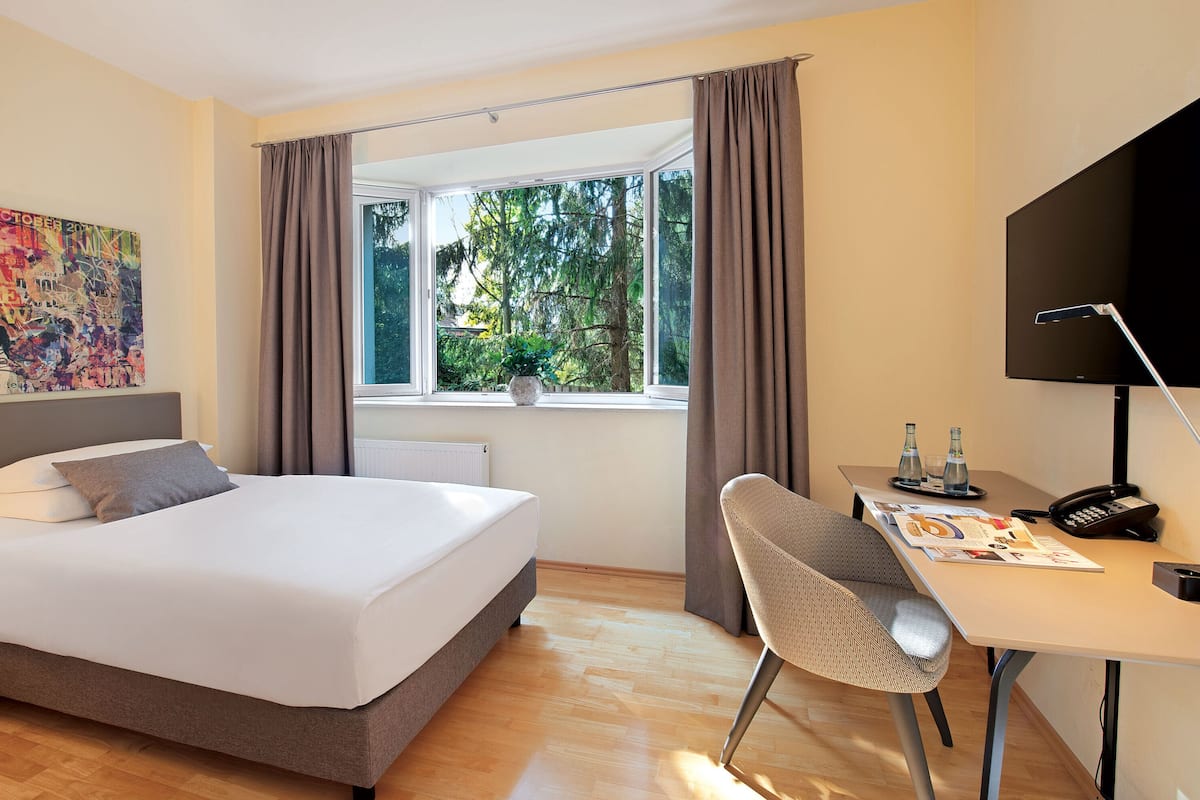 A hotel room with a bed, a desk with a chair, a flat-screen TV, and a window with curtains.