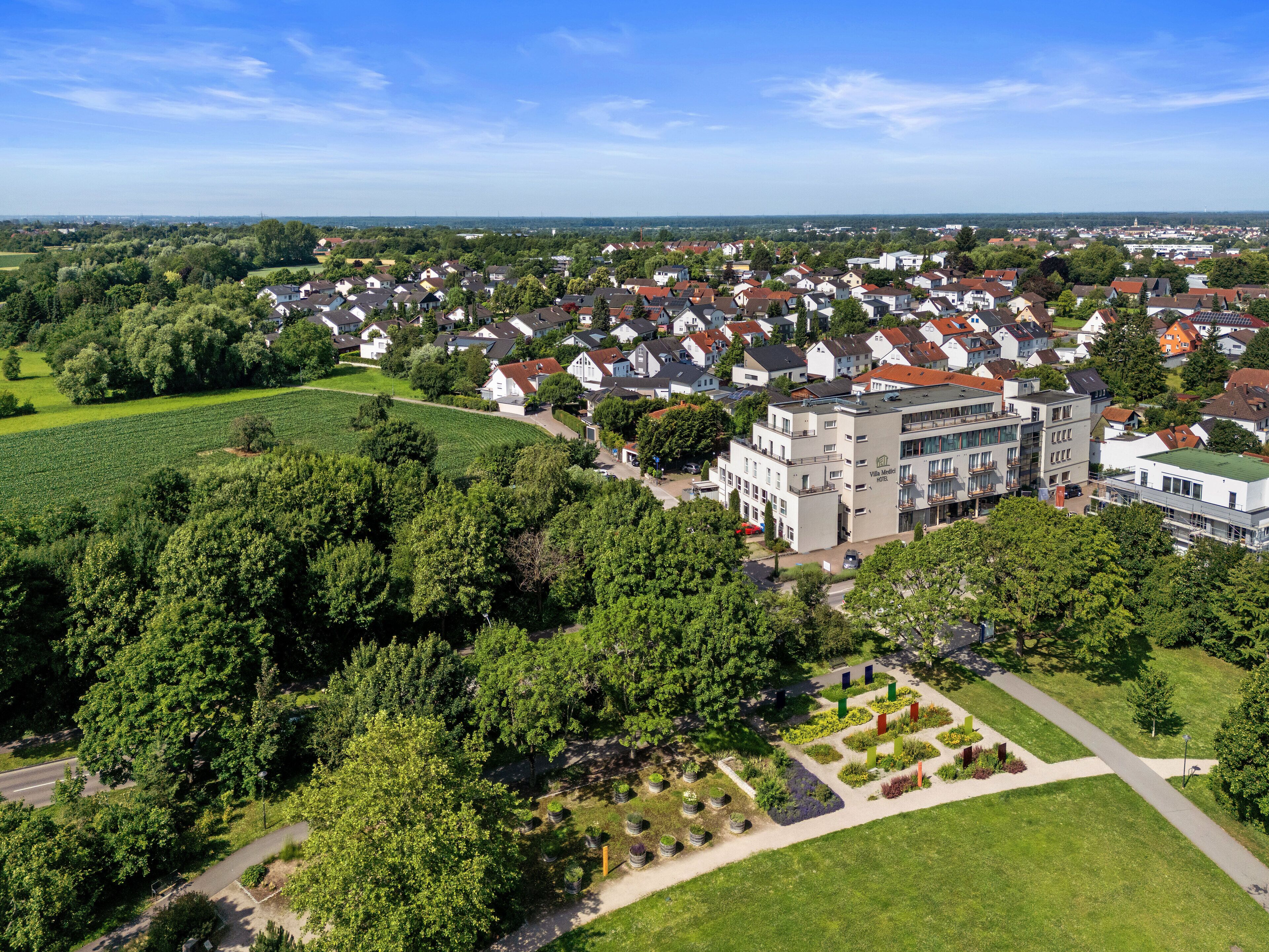An aerial view of a residential area with a prominent hotel building featuring a modern design, surrounded by lush greenery and a well-maintained garden.