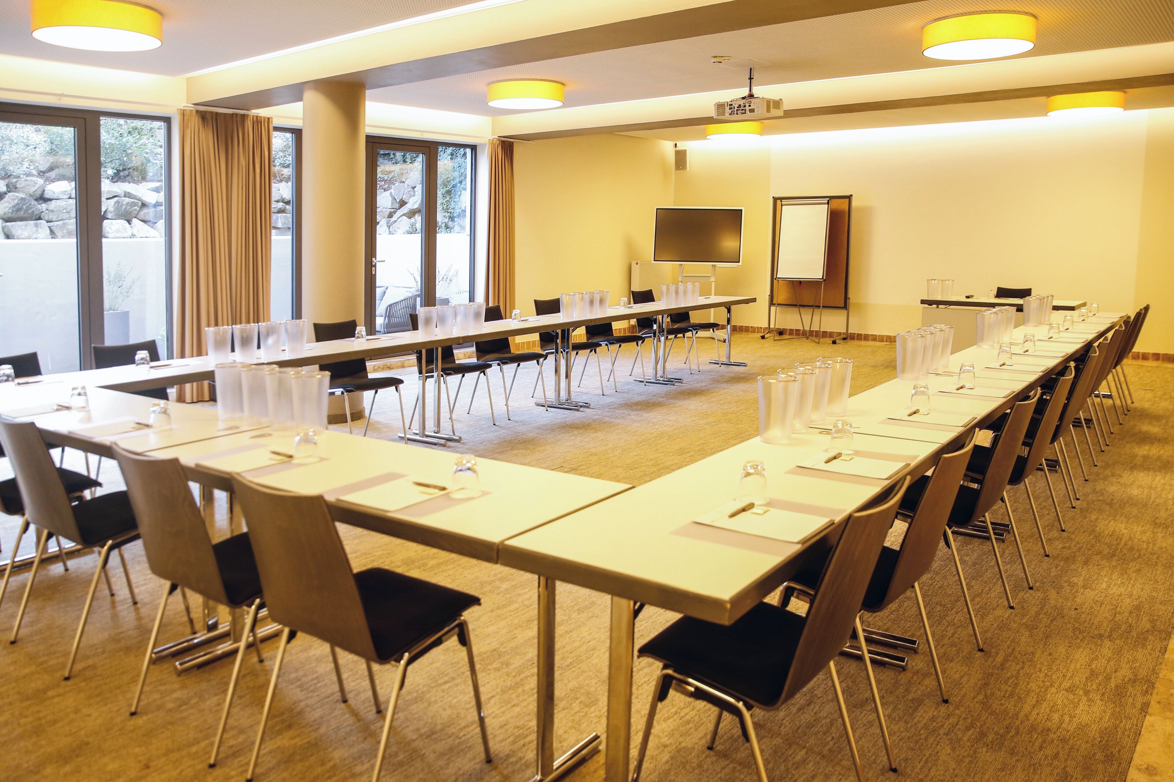 A conference room with long tables, chairs, a projector screen, and a large window with curtains.