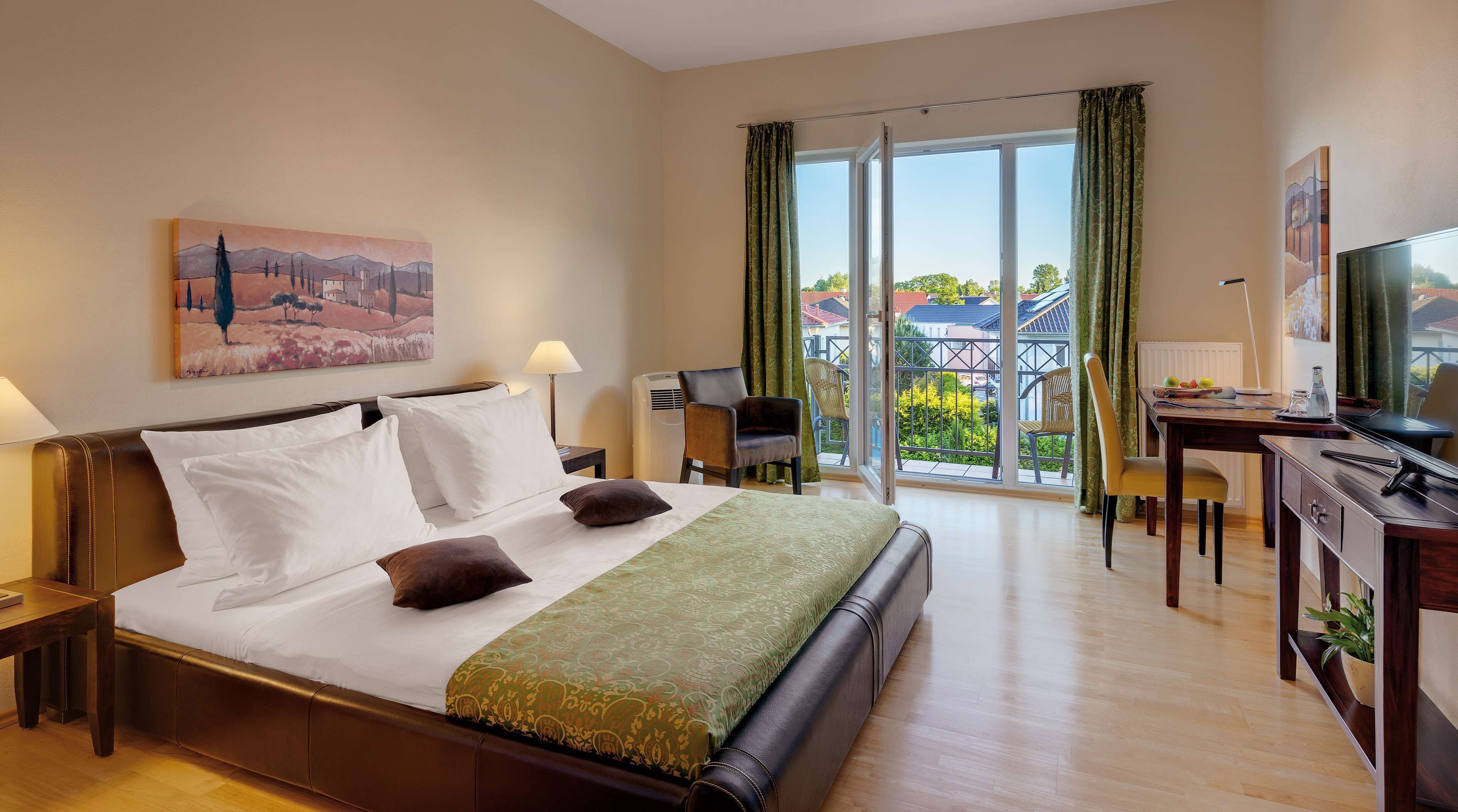 Quarto de hotel com uma cama grande, uma escrivaninha, uma cadeira, uma televisão e uma varanda com vista para edifícios e áreas verdes.