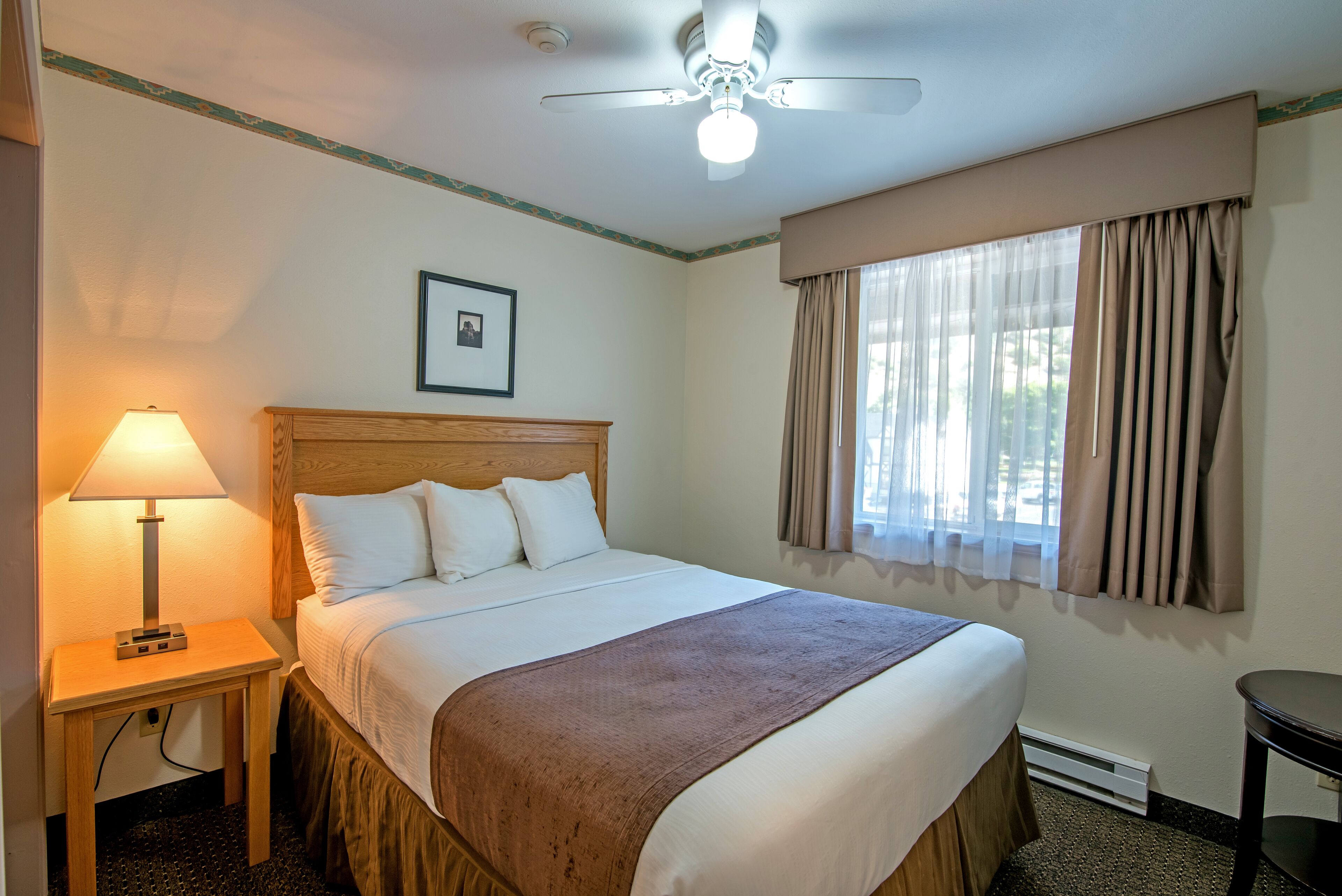A hotel room with a bed, a nightstand with a lamp, a ceiling fan, and a window with curtains.