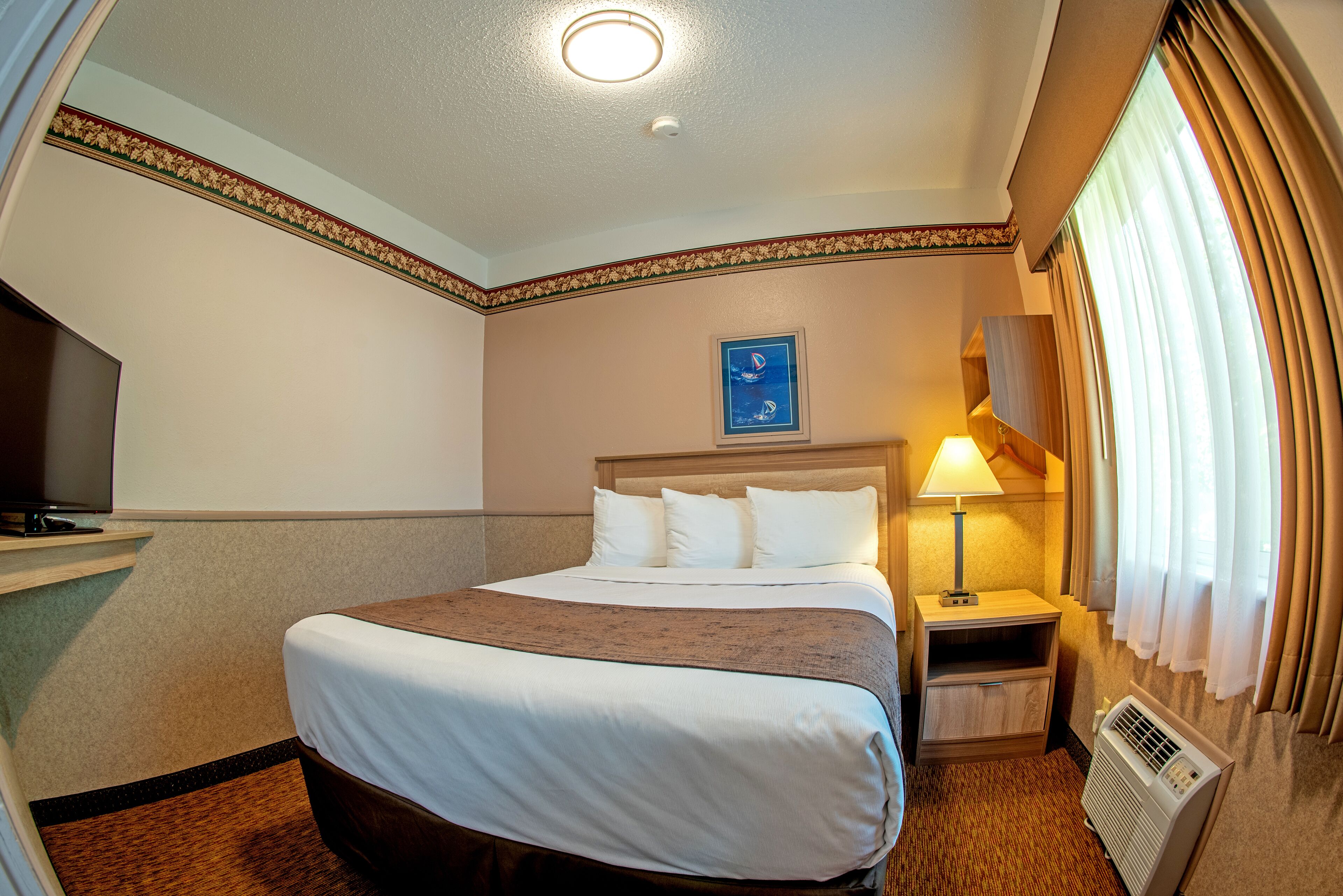 A hotel room with a bed, a nightstand with a lamp, a TV, and a window with curtains.