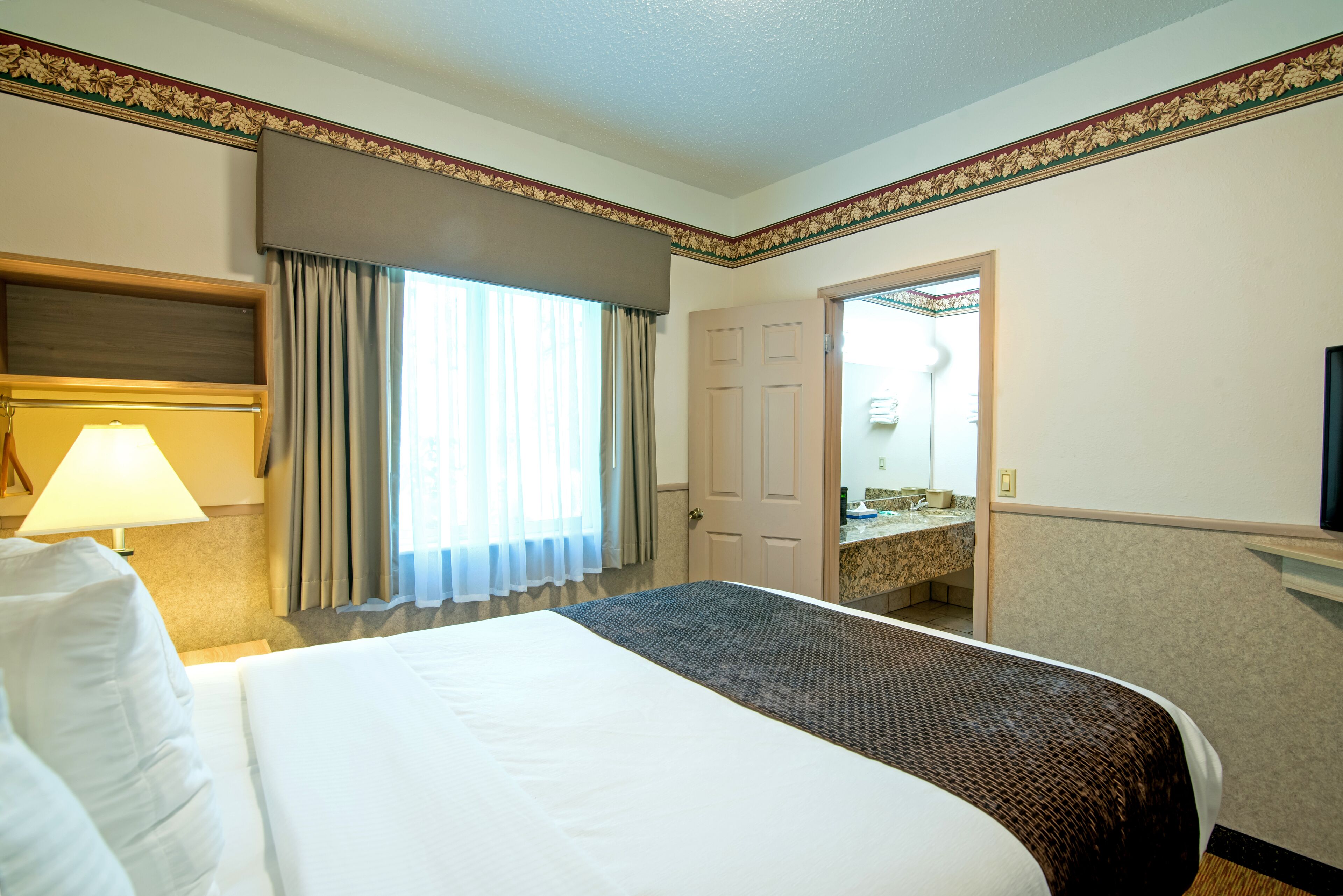 Habitación de hotel con una cama grande, una lámpara de noche, una ventana con cortinas, un baño con lavamanos y espejo, y una puerta que da a otra habitación.