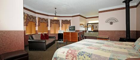A hotel room with a large bed, a sofa, a desk with a television, a fireplace, and a mirror.