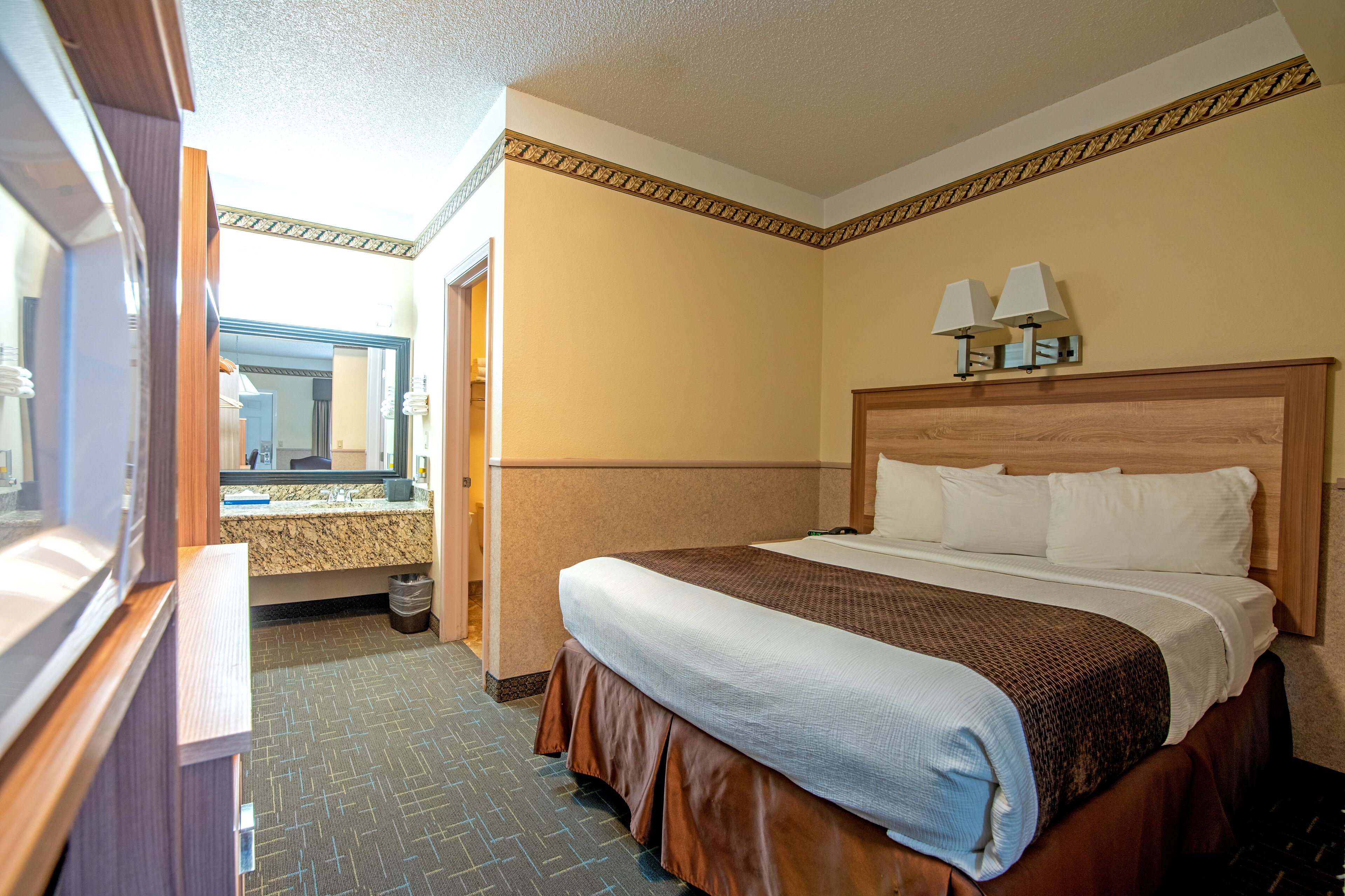 A hotel room with a large bed, a wooden headboard, two bedside lamps, and a bathroom visible through an open door.
