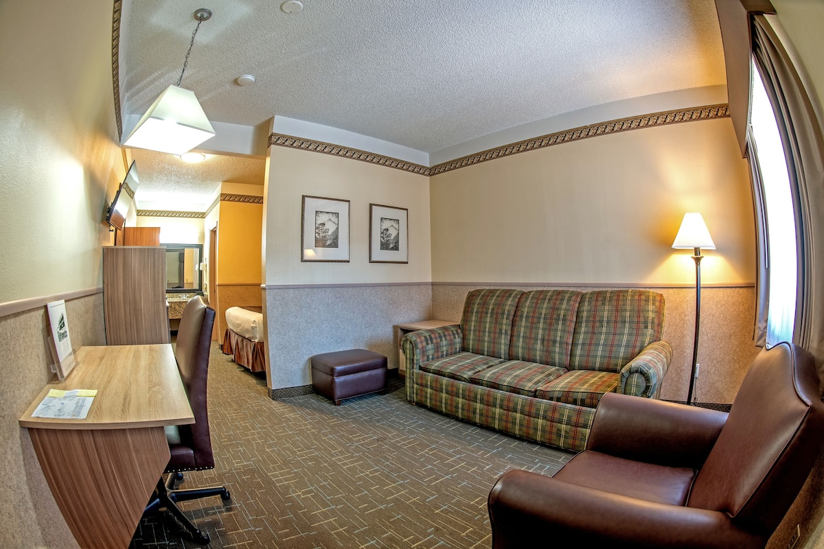 A hotel room with a wooden desk, a plaid sofa, a leather armchair, and a small ottoman. There are two framed pictures on the wall and a lamp on the right side.