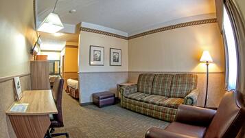 A hotel room with a wooden desk, a plaid sofa, a leather armchair, and a small ottoman. There are two framed pictures on the wall and a lamp on the right side.