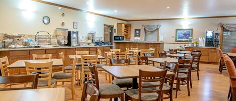 A spacious dining area with multiple wooden tables and chairs, a kitchen area with appliances, and a counter with various food items.