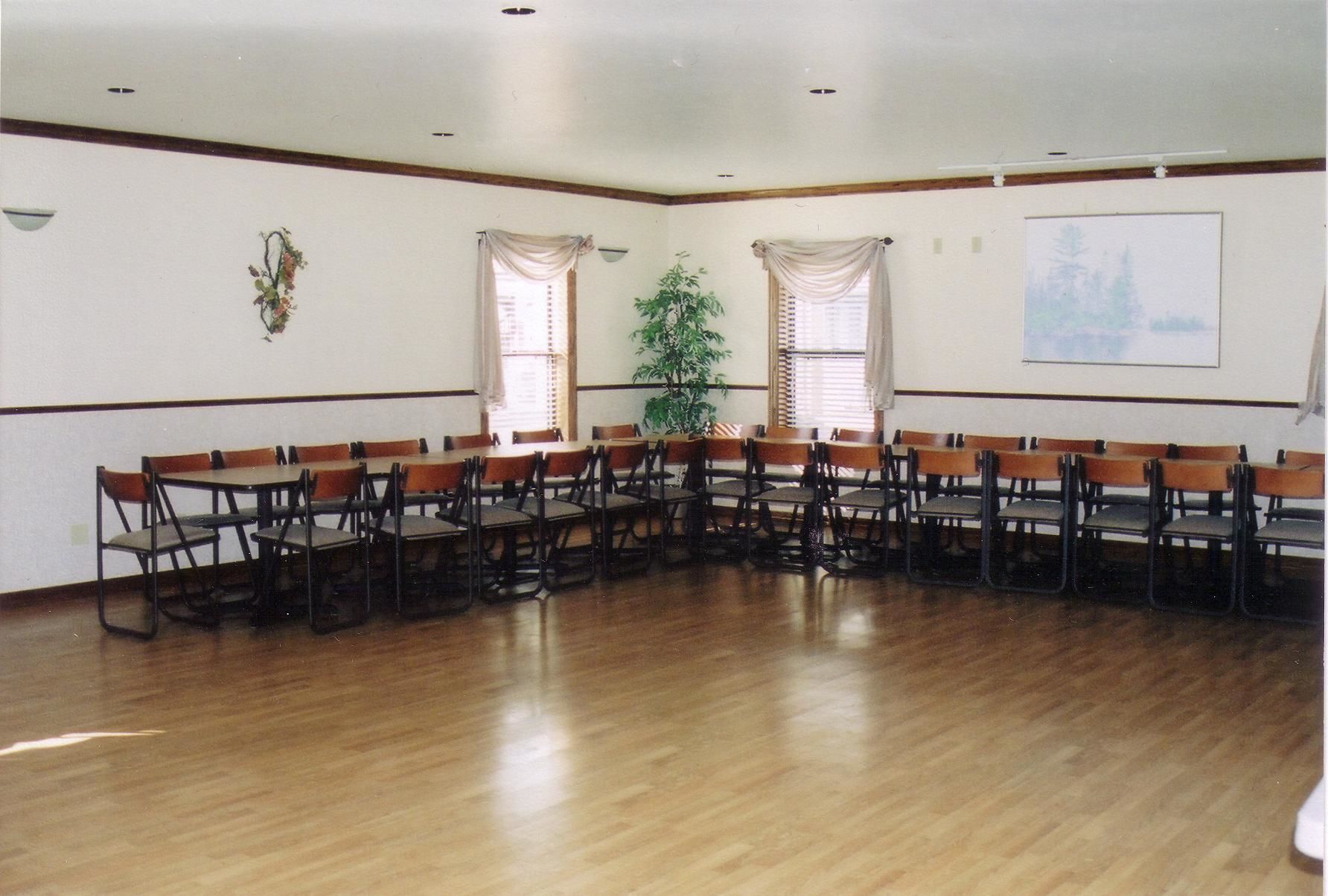 Un salón de conferencias con mesas y sillas de madera dispuestas en forma de U, ventanas amplias con cortinas, una planta en maceta y un cuadro en la pared.