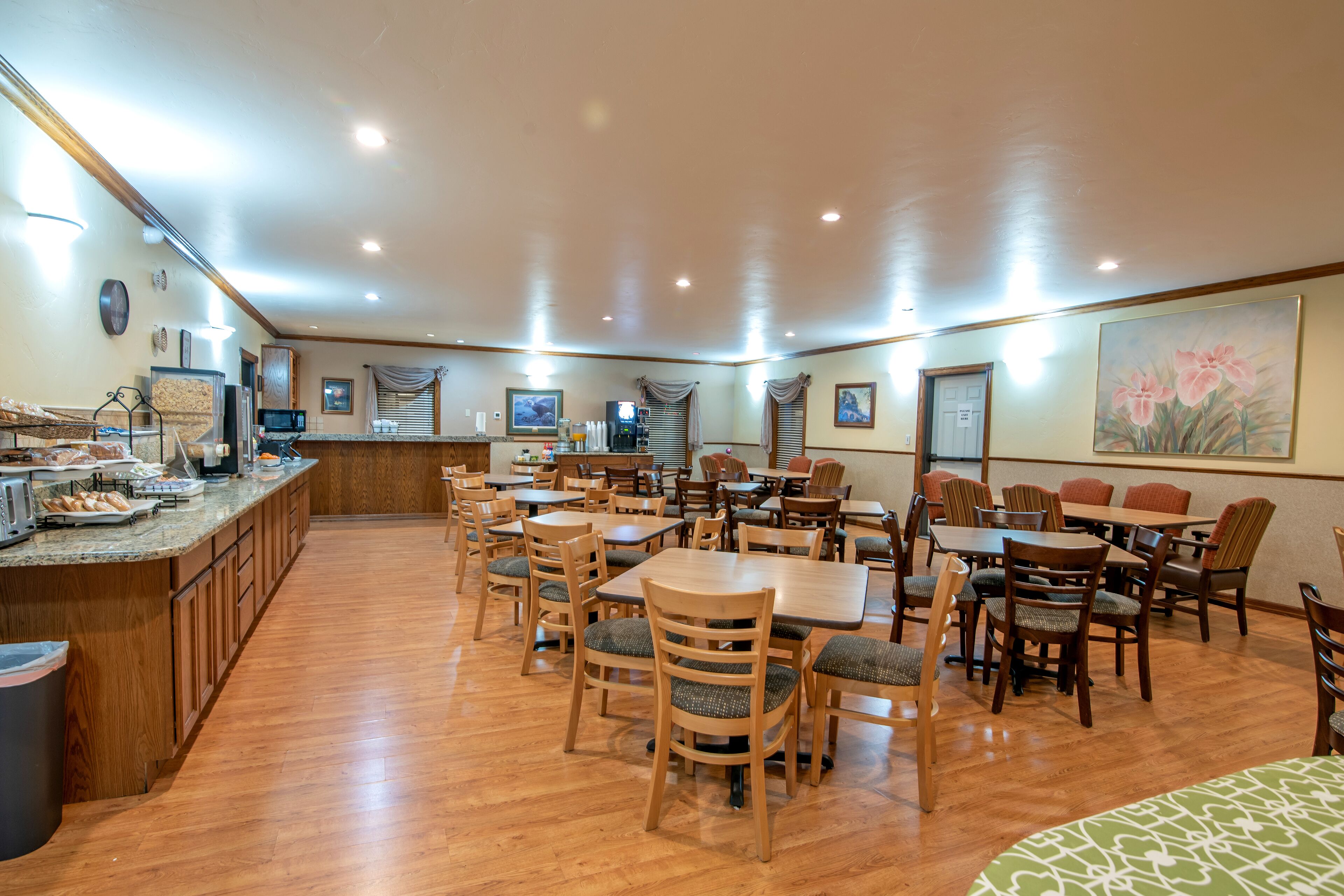 A spacious dining area with wooden tables and chairs, a counter with food items, and a large painting on the wall.