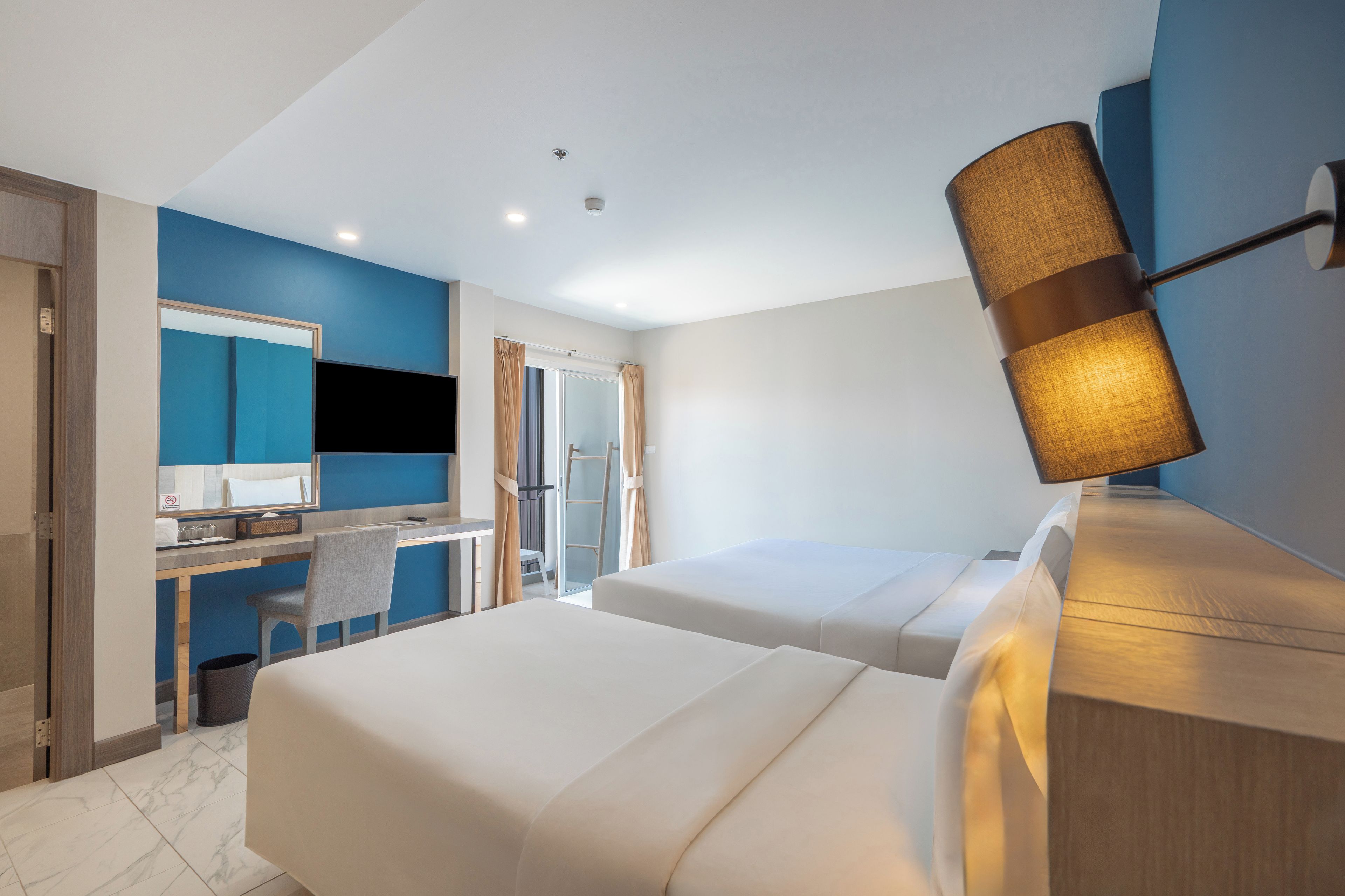 A hotel room with two beds, a desk, a chair, a mirror, and a lamp.